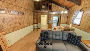 Un intérieur de chalet chaleureux avec des murs en bois, une chambre à coucher située en mezzanine, un coin repas et un canapé agrémenté de coussins.