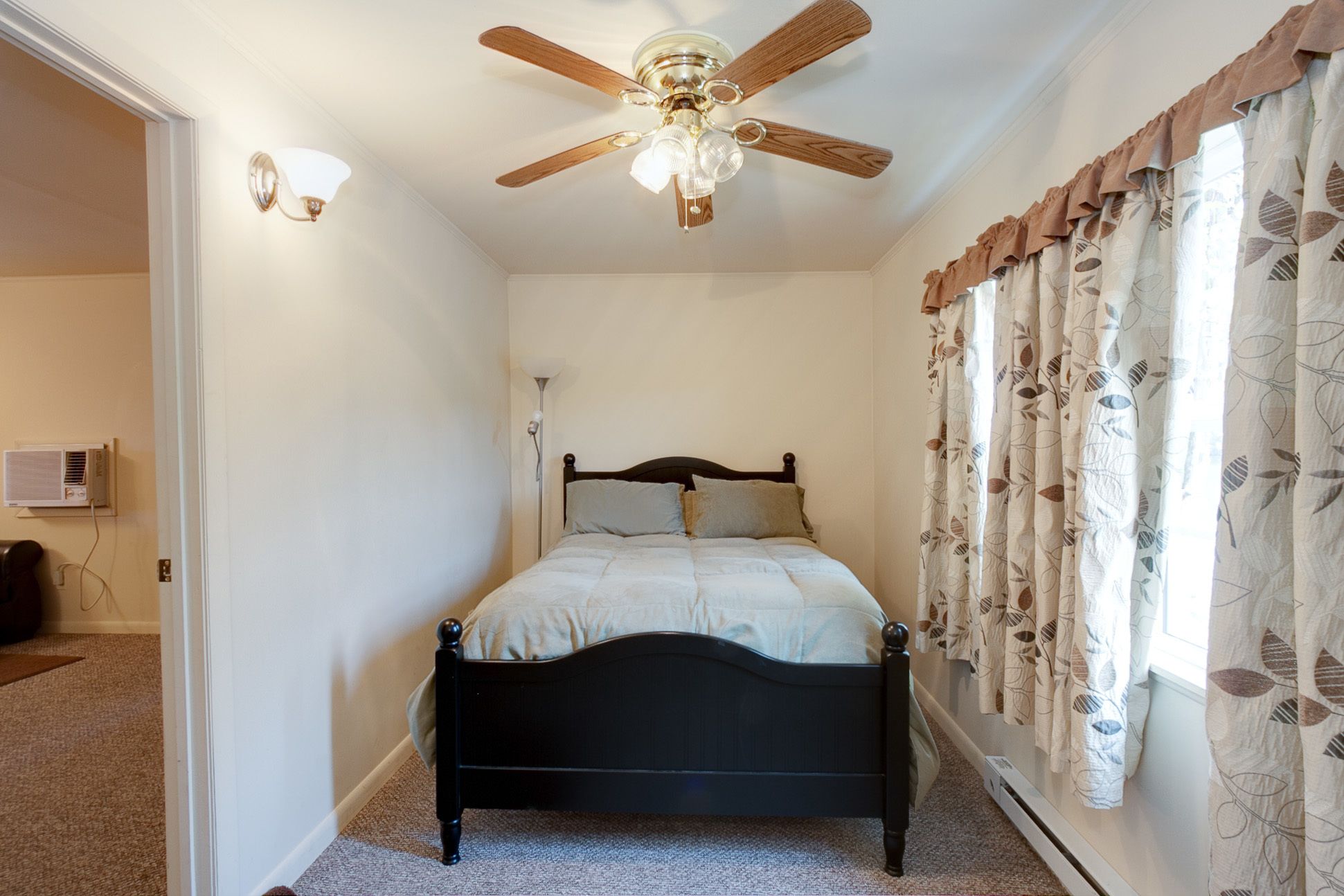 A bedroom with a bed, a ceiling fan, a window with curtains, and a wall-mounted air conditioner.