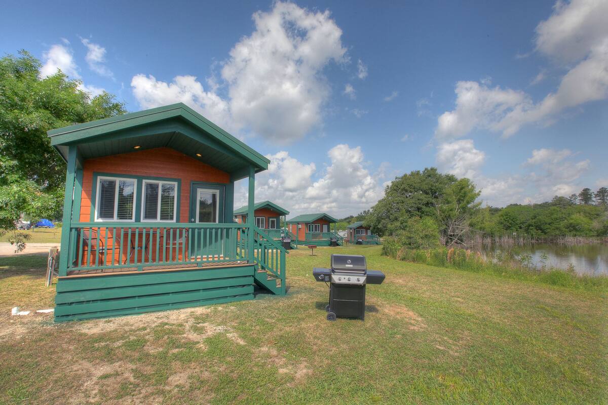 A cabin with a porch and a grill in a grassy area near a body of water.
