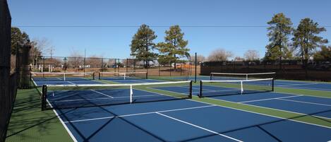 Una cancha de tenis al aire libre con varias canchas, una cerca y árboles al fondo.