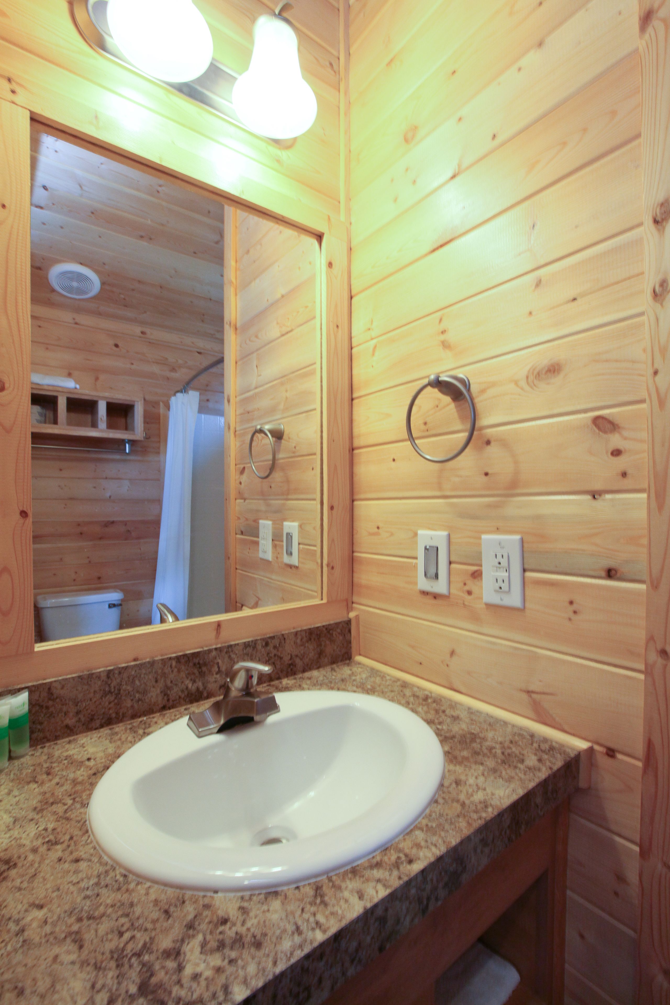 A wooden cabin bathroom with a granite countertop sink, a large mirror, and a ring light above the mirror.