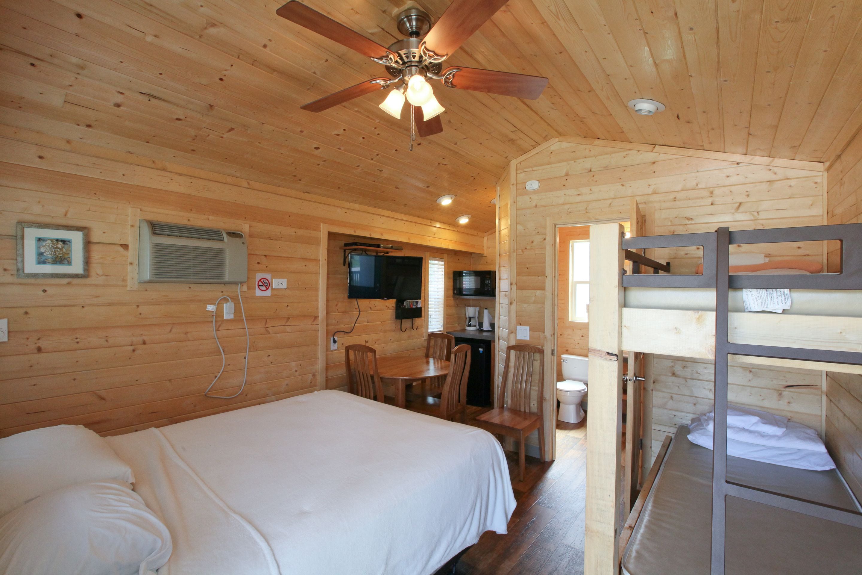 A wooden cabin room with a bed, a desk, a chair, a bunk bed, and a ceiling fan.