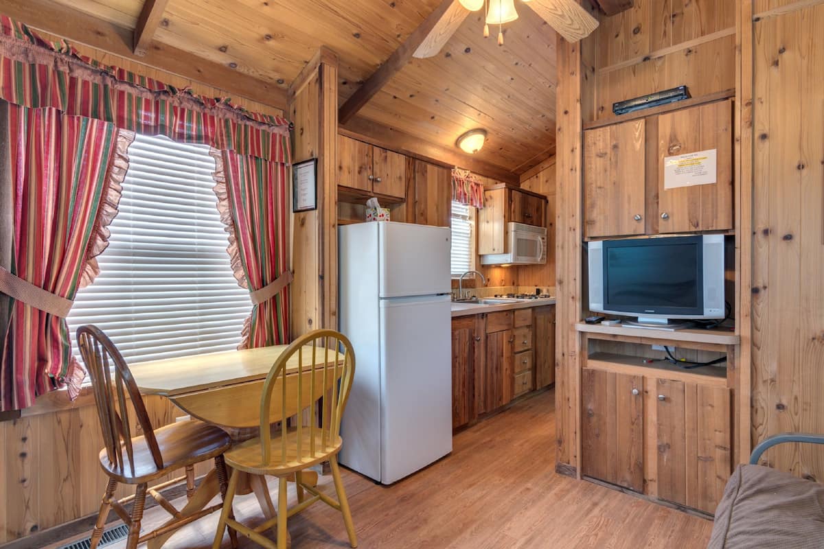 A wooden cabin interior with a dining table, chairs, a refrigerator, a microwave, and a television.