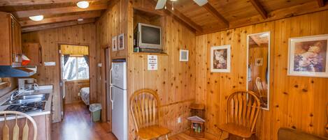 A cabin interior with wooden walls, a ceiling fan, a kitchenette, two wooden chairs, and a couch.