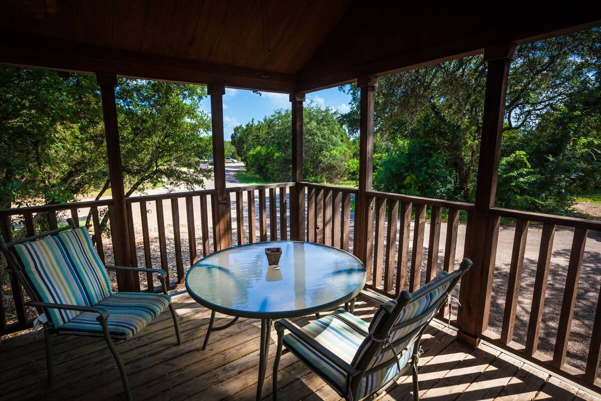 A wooden deck with a glass table and striped chairs, overlooking a road and greenery.