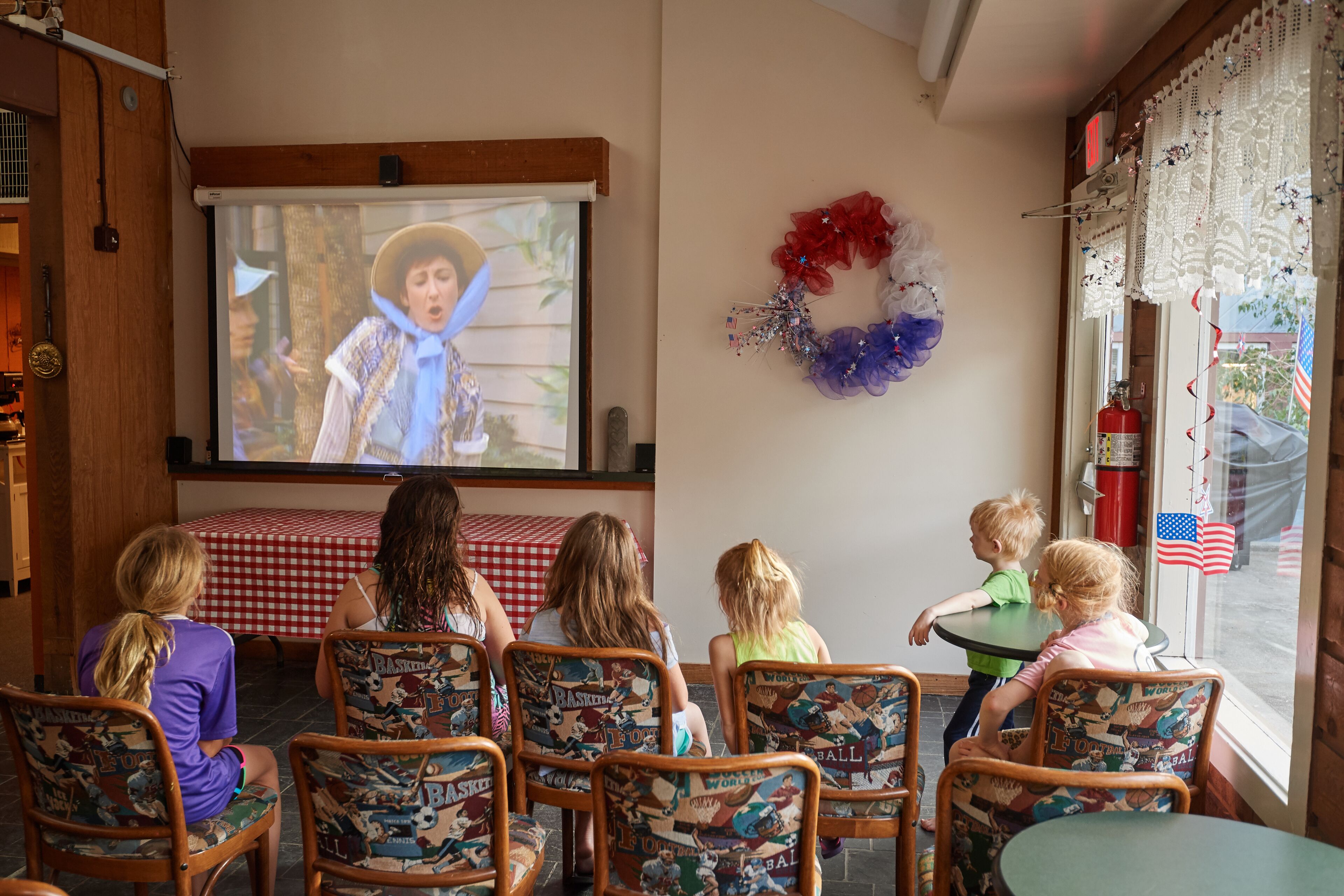 A group of children watching a movie on a projector screen in a room with wooden chairs and a red and white checkered tablecloth.