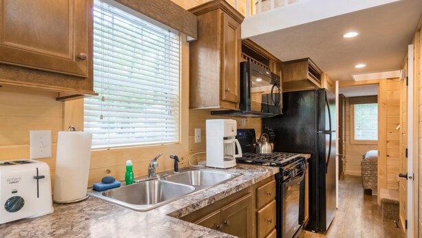 A compact kitchen with wooden cabinets, a stainless steel sink, a black refrigerator, a microwave, and a gas stove. - Williamsburg RV & Camping Resort (Williamsburg)