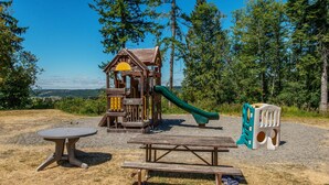 A playground with a wooden playhouse, slide, picnic table, and plastic chairs. - Chehalis RV & Camping Resort (Chehalis)