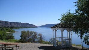 A gazebo with a gravel ground, a picnic table, and a view of a lake with cliffs in the background.