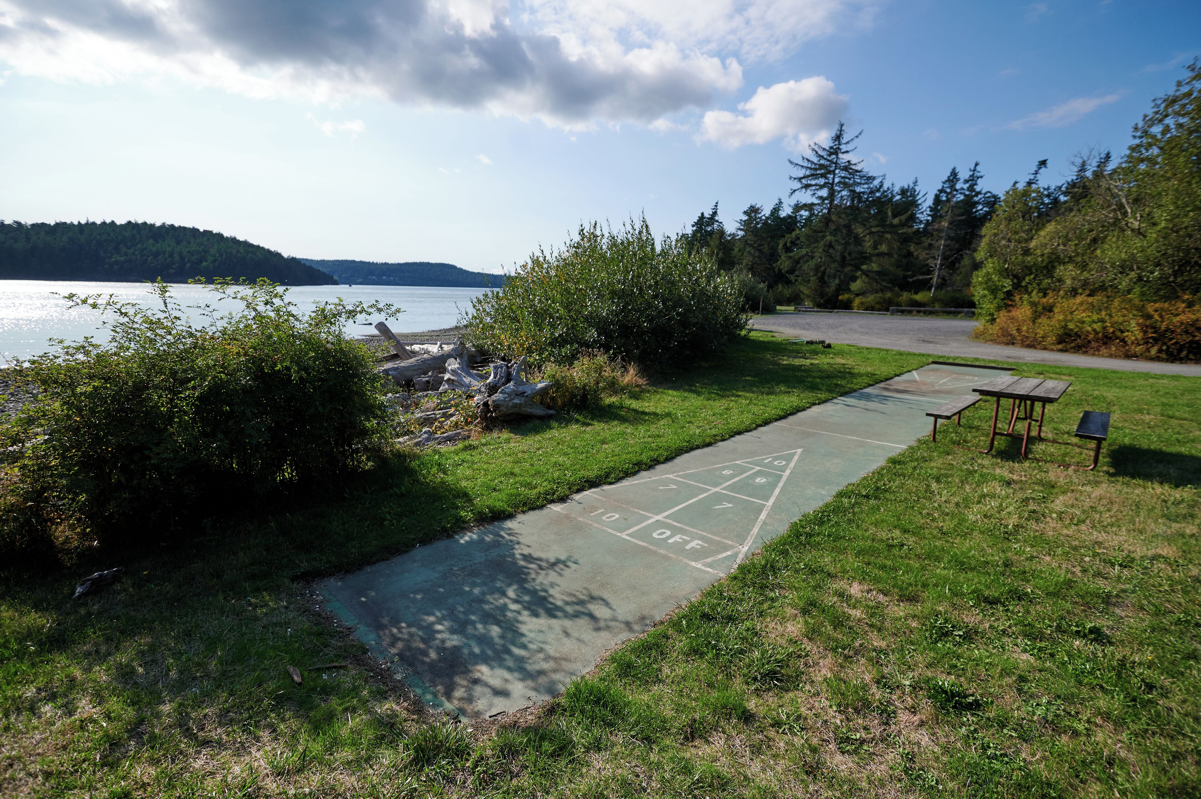A grassy area with a shuffleboard court, a small table, and a bench near a body of water.