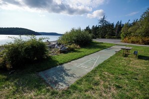 A grassy area with a shuffleboard court, a small table, and a bench near a body of water. - La Conner RV & Camping Resort (La Conner)