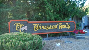 A wooden sign with the text "Thousand Trails" and a logo, surrounded by greenery and a flower pot. - La Conner RV & Camping Resort (La Conner)