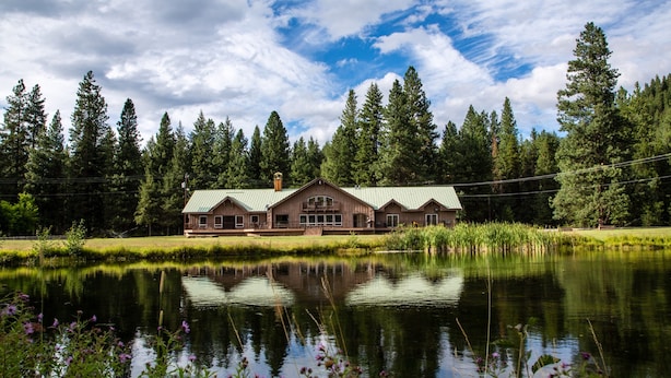 A lodge with a large central building and a smaller structure to the left, surrounded by a pond and dense forest.