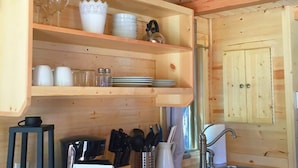 A wooden kitchen with shelves holding dishes, glasses, and utensils. There's a sink with a faucet, a black lantern, and a sign with text.