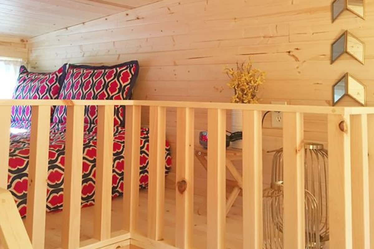 Interior de una cabaña de madera con techo inclinado, baranda de madera y una cama con ropa de cama estampada.