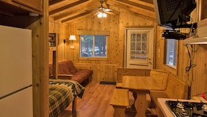 A cozy cabin interior with a wooden ceiling, a bed, a dining table, a stove, and a television mounted on the wall.