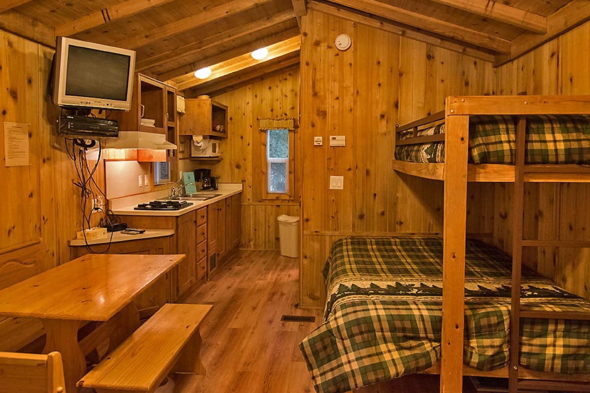 Una cabaña con kitchenette, una cama individual con manta escocesa y una mesa de comedor de madera.