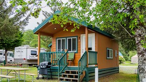 A small wooden cabin with a porch, a grill, and picnic tables. - Long Beach RV & Camping Resort (Ilwaco)