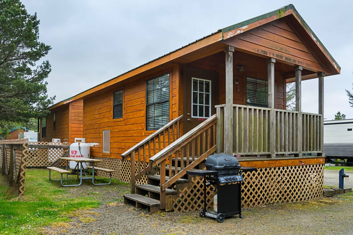 A wooden cabin with a porch, a grill, picnic tables, and a trash can.