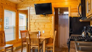 A wooden cabin interior with a dining table, chairs, a TV mounted on the wall, a refrigerator, and a stove. - Long Beach RV & Camping Resort (Ilwaco)