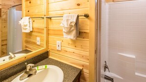 A bathroom with wooden walls, a granite countertop, a white sink, a mirror, and a shower area. - Long Beach RV & Camping Resort (Ilwaco)
