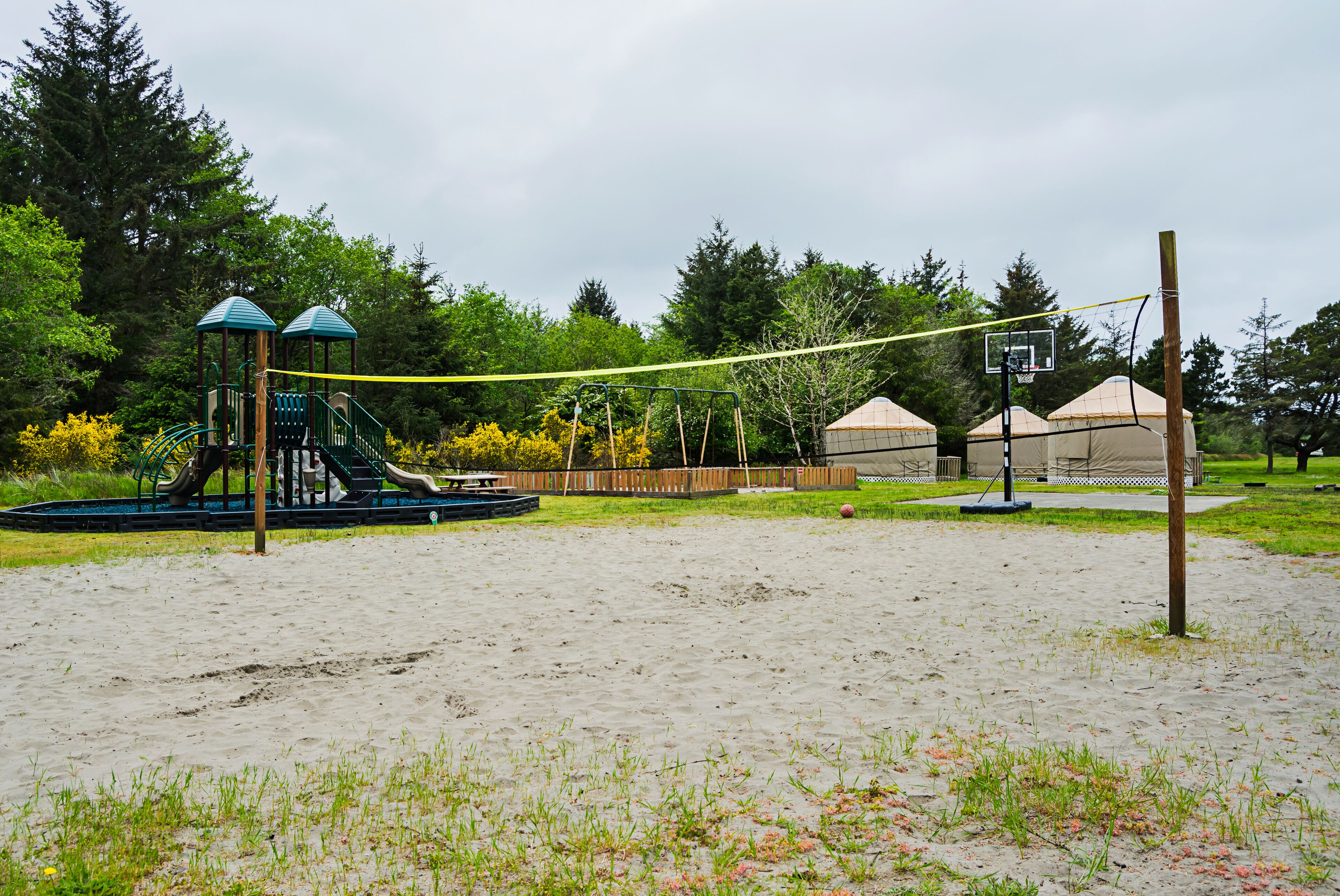 A playground with a slide and climbing structures, a basketball hoop, and a sandy area.