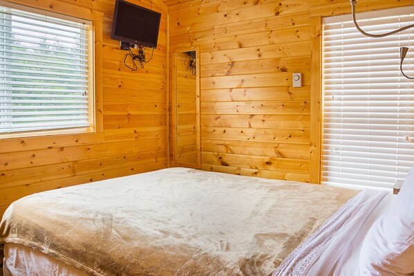 A wooden cabin room with a bed, a television mounted on the wall, and a window with blinds.