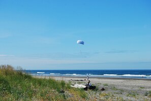 A beach with a parasail in the distance, a person sitting on the sand, and the ocean.