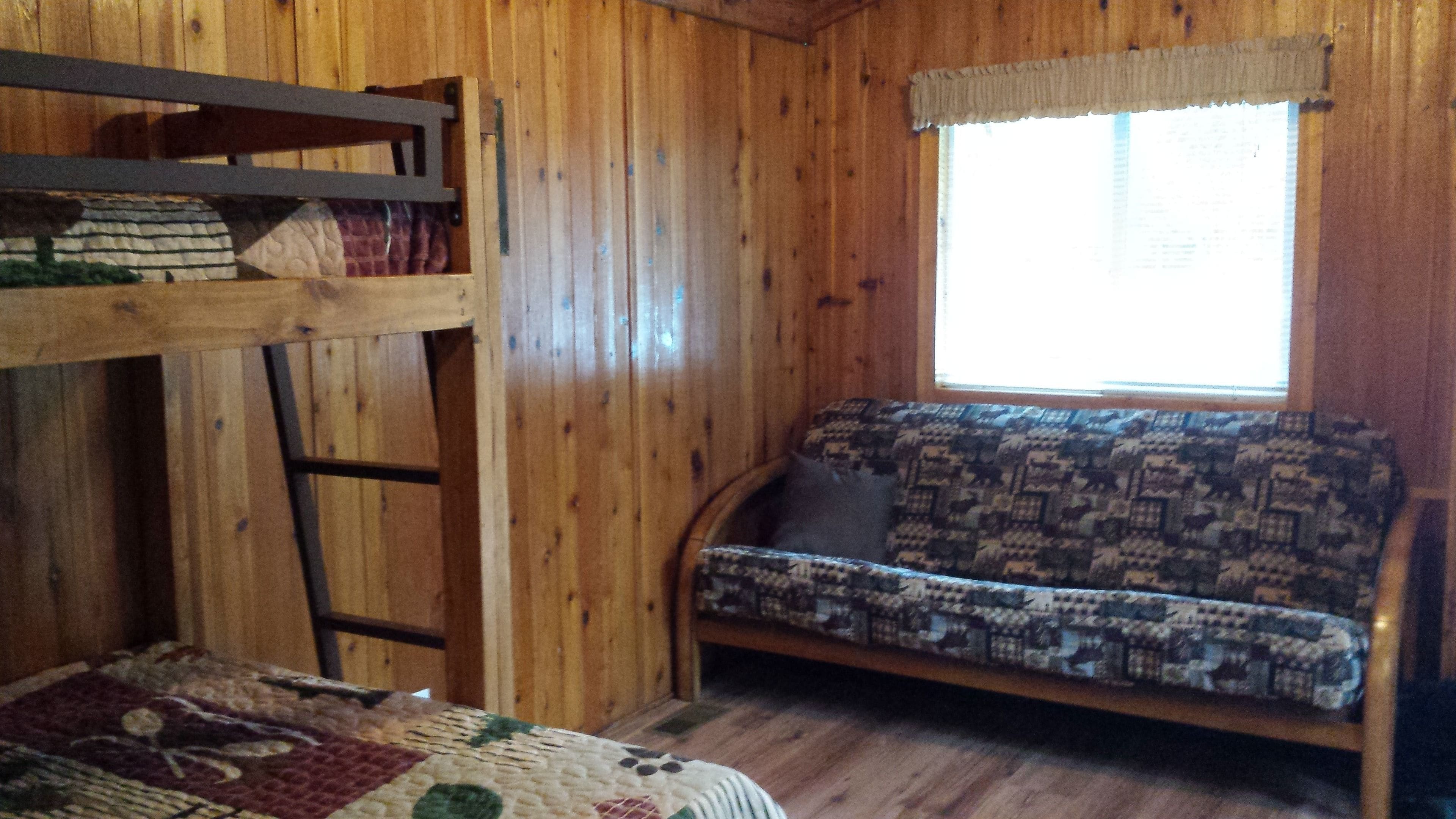 A wooden cabin room with a bunk bed, a sofa with patterned cushions, and a window with a sheer curtain.