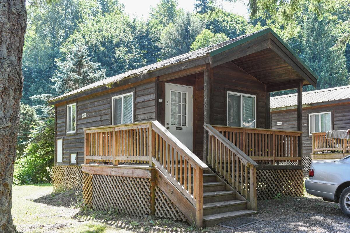 A rustic cabin with a wooden deck, a front porch, and a car parked nearby.