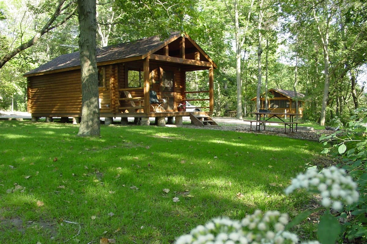 A rustic wooden cabin with a porch, surrounded by a grassy area and trees.