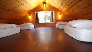 Un dormitorio acogedor en el ático, con paredes de madera y techo inclinado. Hay dos camas individuales y una cama más grande. Una ventana con cortinas deja entrar luz natural.