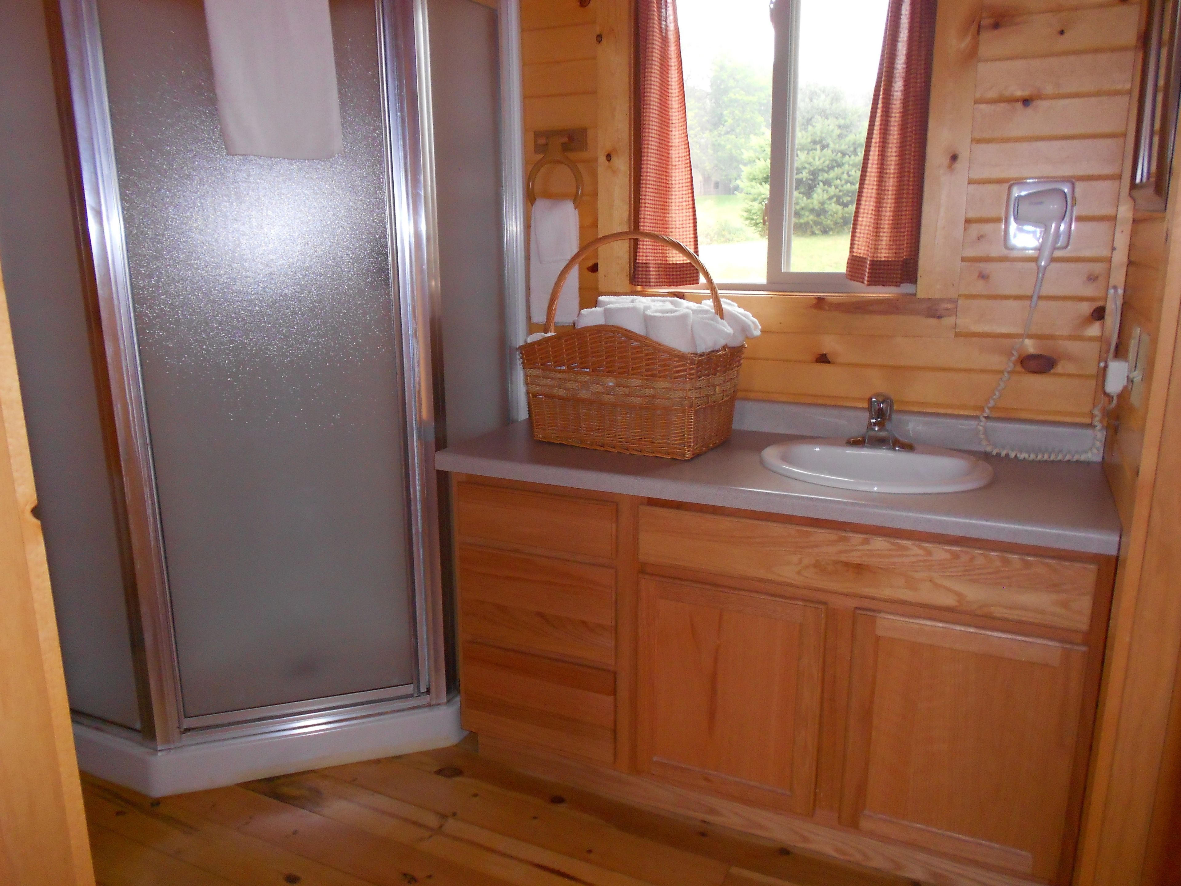 Un baño de cabaña de madera con mampara de vidrio, un lavamanos, una cesta de mimbre con toallas y una ventana con cortinas.