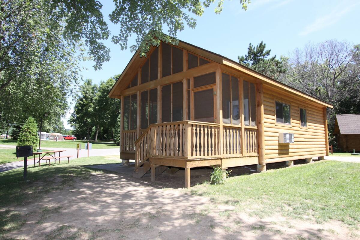 A wooden cabin with a large windowed porch, surrounded by trees and a picnic table.