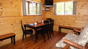 Interior de una cabaña de madera con una mesa de comedor, sillas, un banco y un televisor montado en la pared.