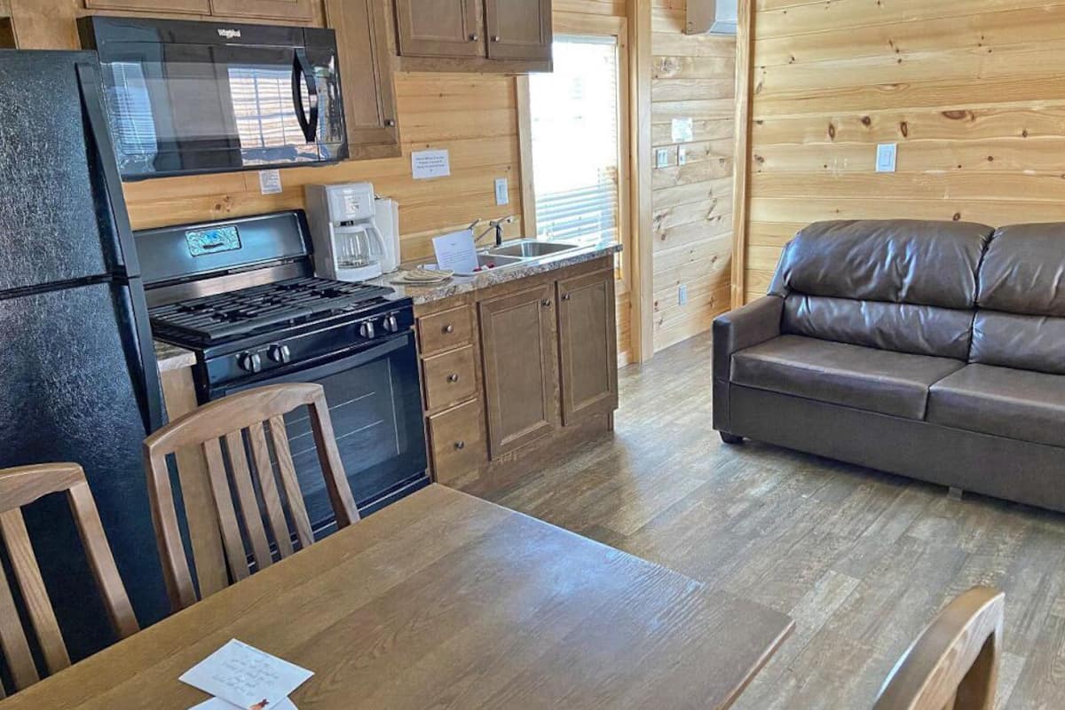 A compact kitchen with a stove, microwave, and coffee maker. There's a wooden dining table with chairs and a leather sofa in a cabin-style room.