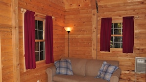 A wooden cabin room with a brown sofa, two plaid pillows, and two windows with red curtains. - Tranquil Timbers Camping Resort (Sturgeon Bay)