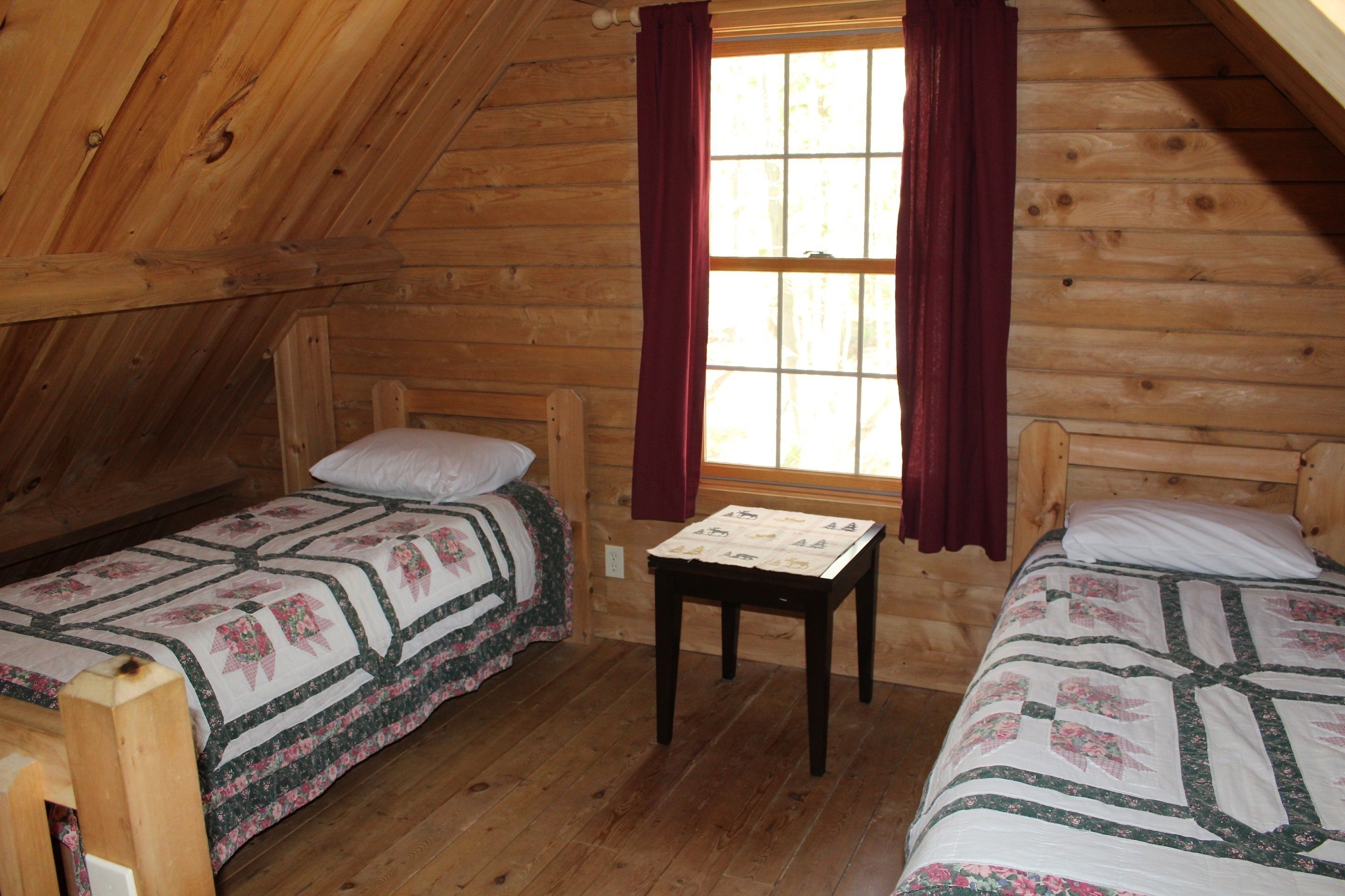 A cozy cabin room with two wooden beds, a small table, and a window with curtains.
