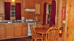 A wooden cabin kitchen with a stove, sink, and dining table with chairs. - Tranquil Timbers Camping Resort (Sturgeon Bay)