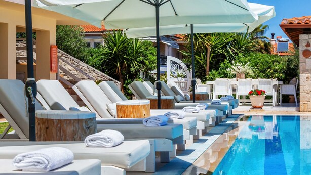 Uma área à beira da piscina com espreguiçadeiras e guarda-sóis, piscina cristalina e um muro de pedra com uma placa.