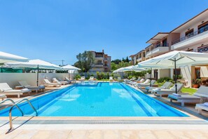 En swimmingpool med liggestole og parasoller, omgivet af bygninger.