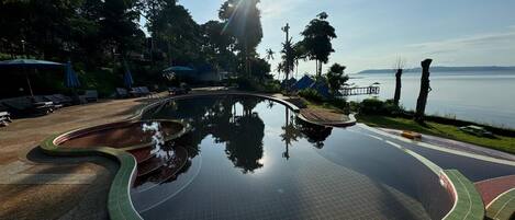 A swimming pool with a curved design, surrounded by lounge chairs and umbrellas, with a view of the sea and trees in the background.
