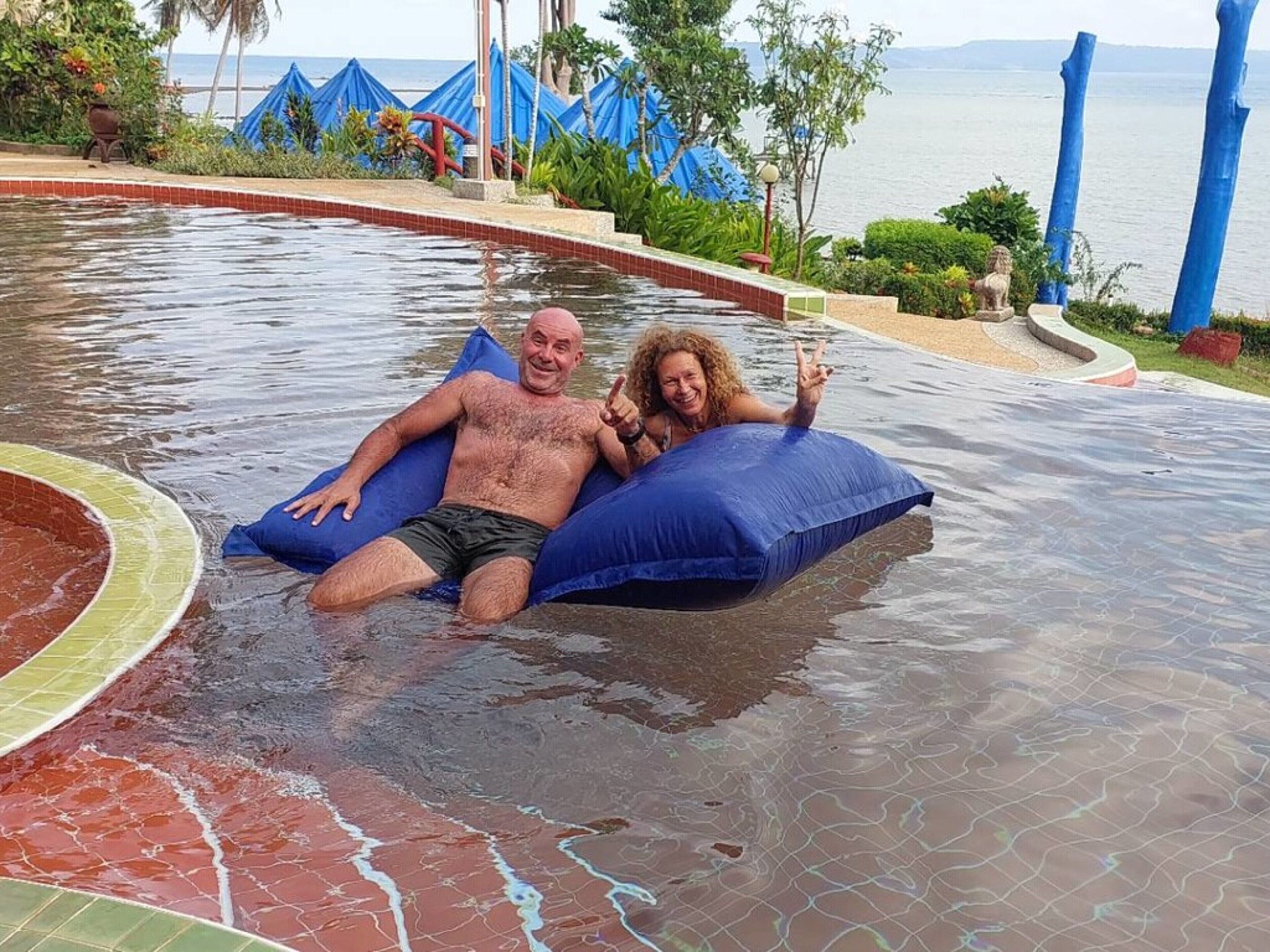 A man and a woman floating on a blue inflatable in a pool with a view of the sea and blue umbrellas in the background.
