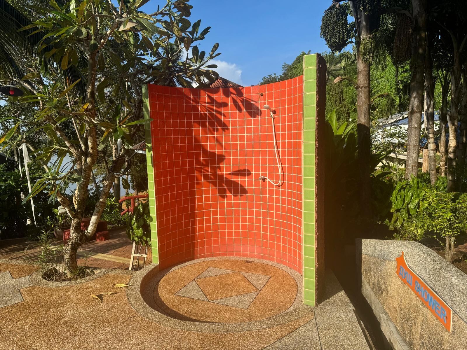 A red-tiled shower enclosure with a green border, surrounded by tropical plants and a circular stone pattern on the ground.