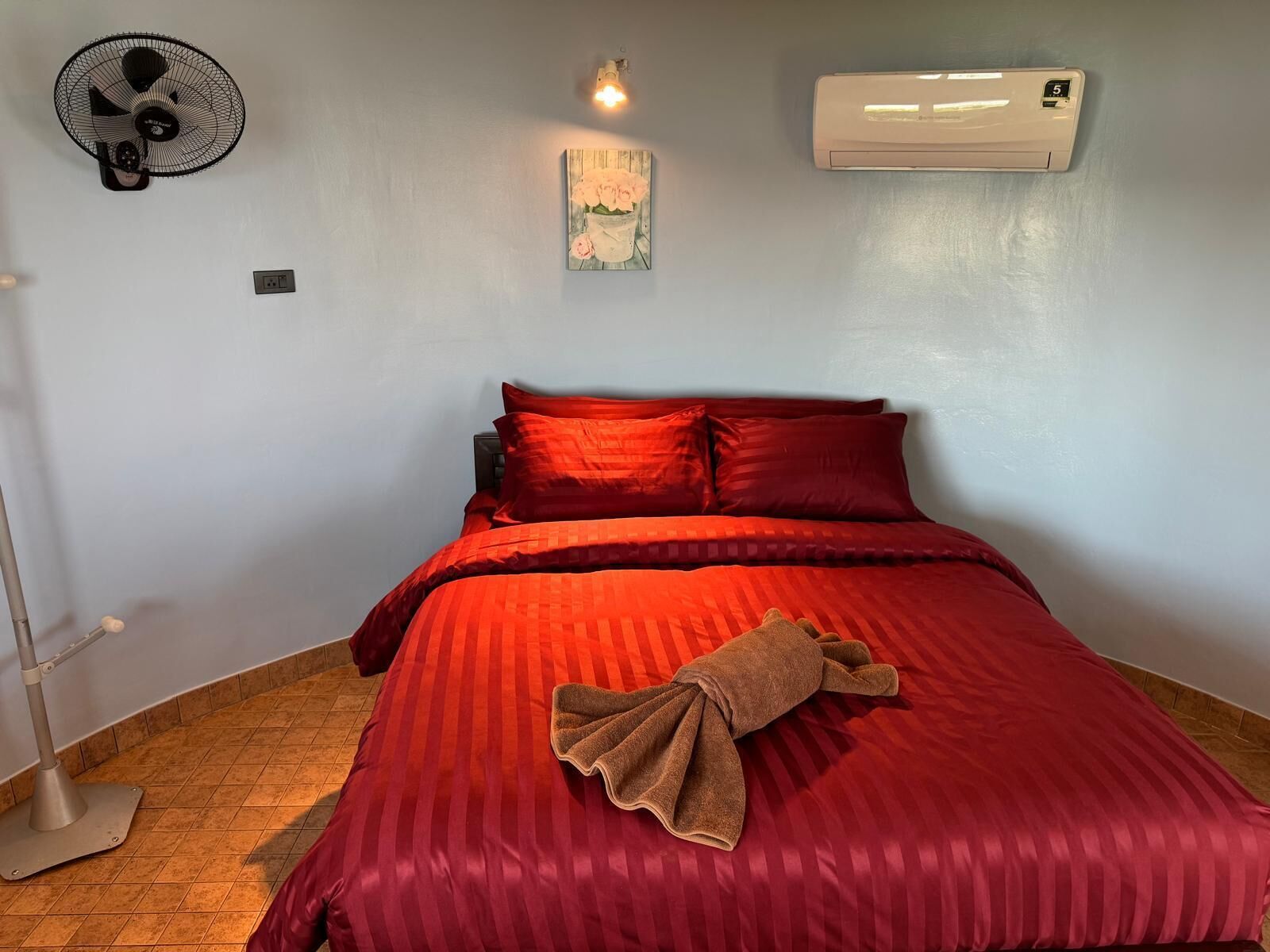A bedroom with a bed, a wall-mounted air conditioner, a wall fan, and a small wall-mounted light.