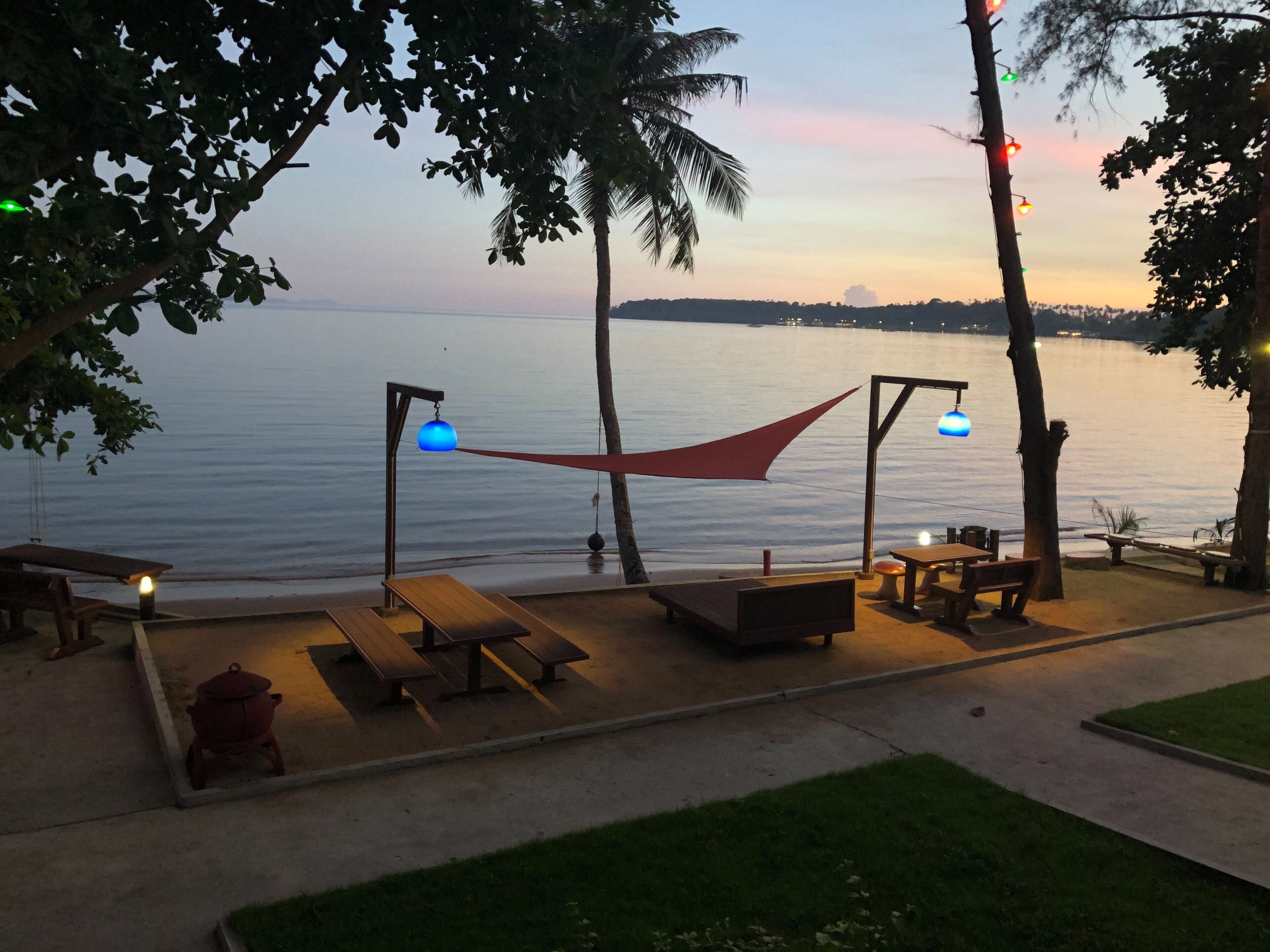 An outdoor seating area with wooden tables and benches, a hammock, and a view of the water at sunset.
