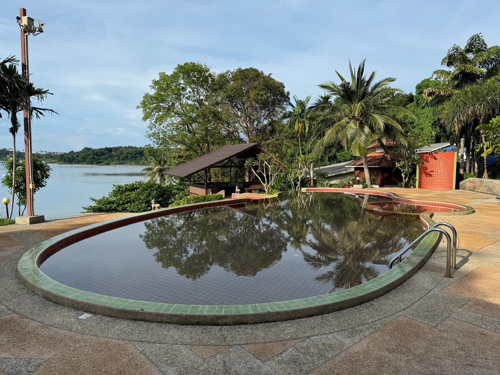 A swimming pool with a tiled edge, surrounded by a paved area with a gazebo and palm trees.