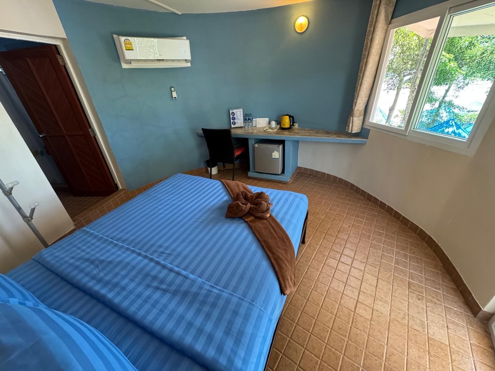 A room with a bed, a desk, a chair, a small refrigerator, and a window with a view of trees.
