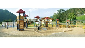 A playground with multiple play structures, including slides and climbing areas, surrounded by a fence and set against a backdrop of mountains and trees. - Hotel-Restaurant Rosengarten (Zell am Ziller)