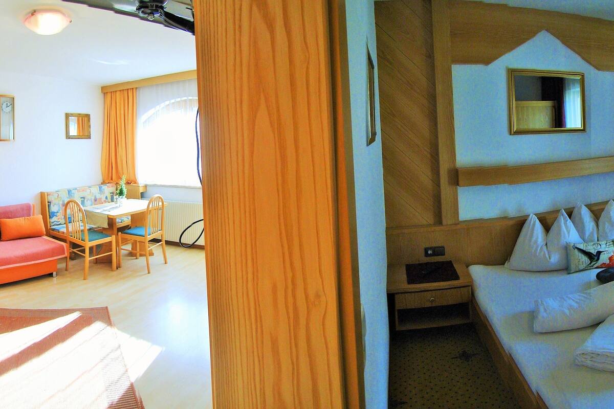Una habitación con comedor, un sofá, una cama y una mesita de noche con lámpara.